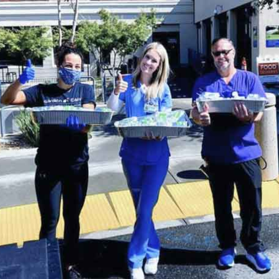 Three Garden of Eat'n staff members, two women and one man, holding catering trays and giving thumbs up.