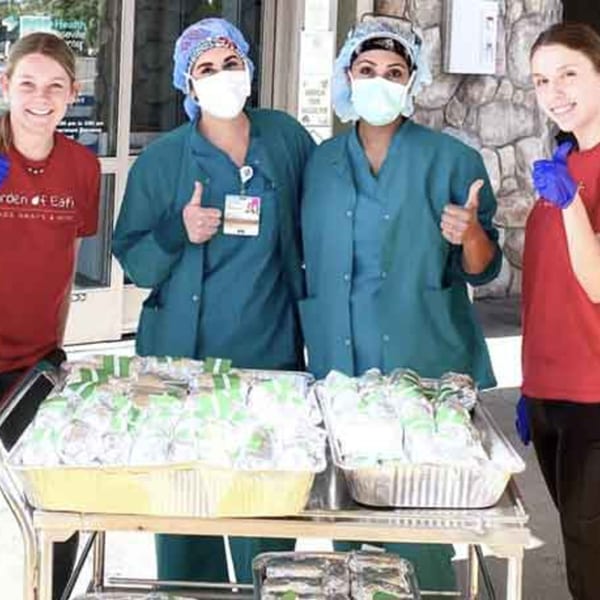 Garden of Eat'n staff and healthcare workers with thumbs up next to trays of packaged meals.