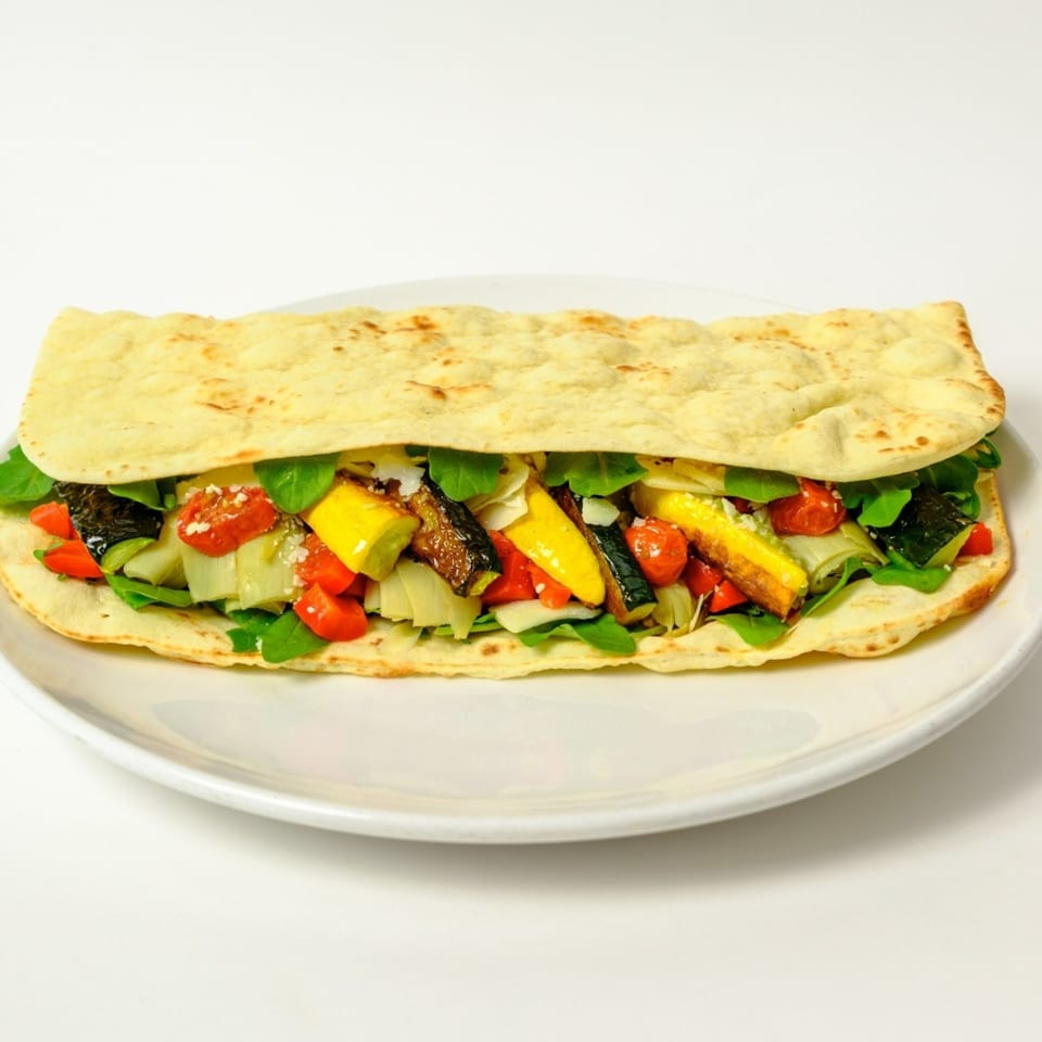 A Garden of Eat'n flatbread sandwich filled with roasted vegetables, greens, and cheese on a white plate.