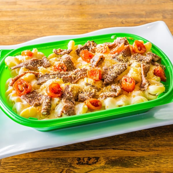 Macaroni and cheese with sliced steak, cherry tomatoes, and breadcrumbs in a green container.