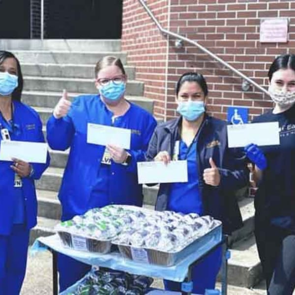 Four masked healthcare workers in blue scrubs stand with trays of food from Garden of Eat'n.